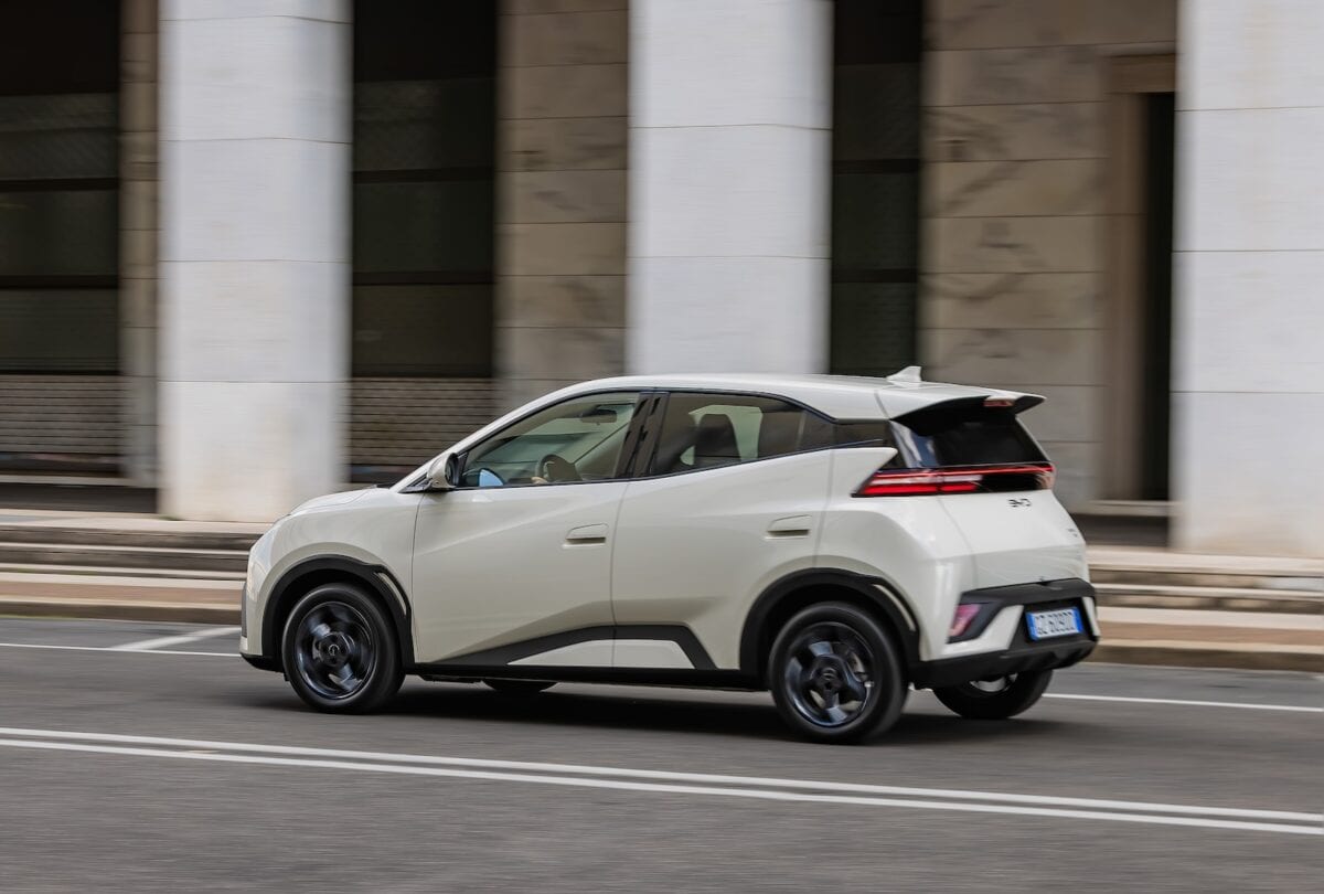 BYD Dolphin Surf, crossover compatto elettrico, fotografato di profilo mentre percorre una strada urbana con colonne moderne sullo sfondo.