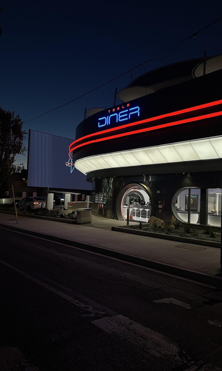 Ingresso del Tesla Diner di Hollywood illuminato di notte con insegna al neon blu e rossa, colonnine Supercharger visibili