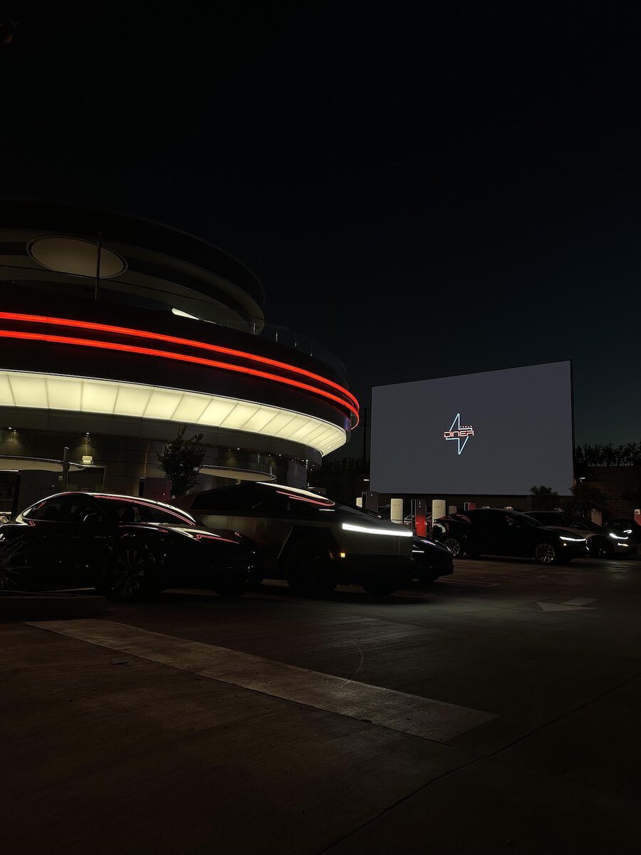 Area parcheggio del Tesla Diner di notte con diverse auto Tesla e Cybertruck, schermo drive-in acceso sullo sfondo