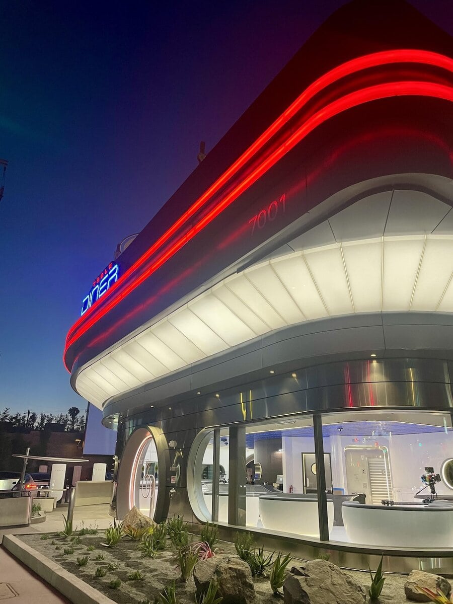 Vista angolare del Tesla Diner con insegna luminosa e vetrate sul lounge interno, colonnine di ricarica visibili a sinistra