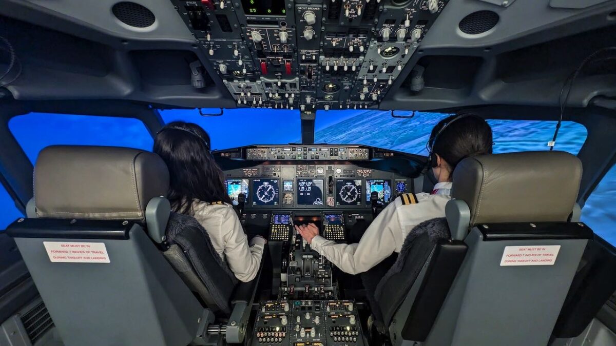 Due aspiranti piloti seduti nella cabina di un simulatore di volo, circondati da comandi realistici e schermi digitali, con vista virtuale sull'oceano attraverso i finestrini.