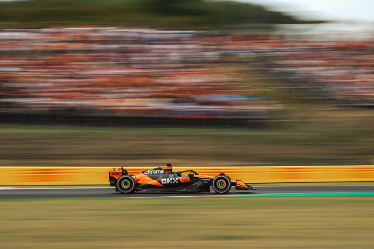 La McLaren Formula 1 2025 di Norris in livrea nero-arancione con sponsor Chrome sfreccia in pista durante una gara, con le tribune gremite sullo sfondo in movimento sfocato.