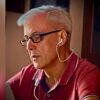 Appassionato di tecnologia e automotive, uomo anziano con occhiali e giacca rossa ascolta musica con auricolari durante una conversazione online.