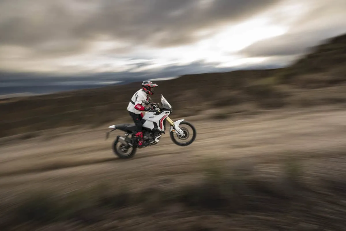 Ducati DesertX 2026 su terreno sterrato, pilota in abbigliamento da off-road, movimento sfocato.